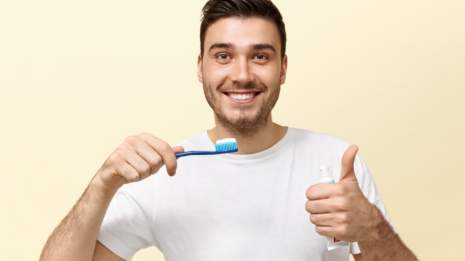 Happy energetic young European guy with stubble holding tooth brush with whitening paste and showing thumbs up gesture being in good mood. Dental care, oral cavity hygiene and healthy teeth concept