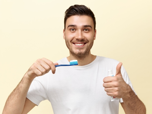 Happy energetic young European guy with stubble holding tooth brush with whitening paste and showing thumbs up gesture being in good mood. Dental care, oral cavity hygiene and healthy teeth concept